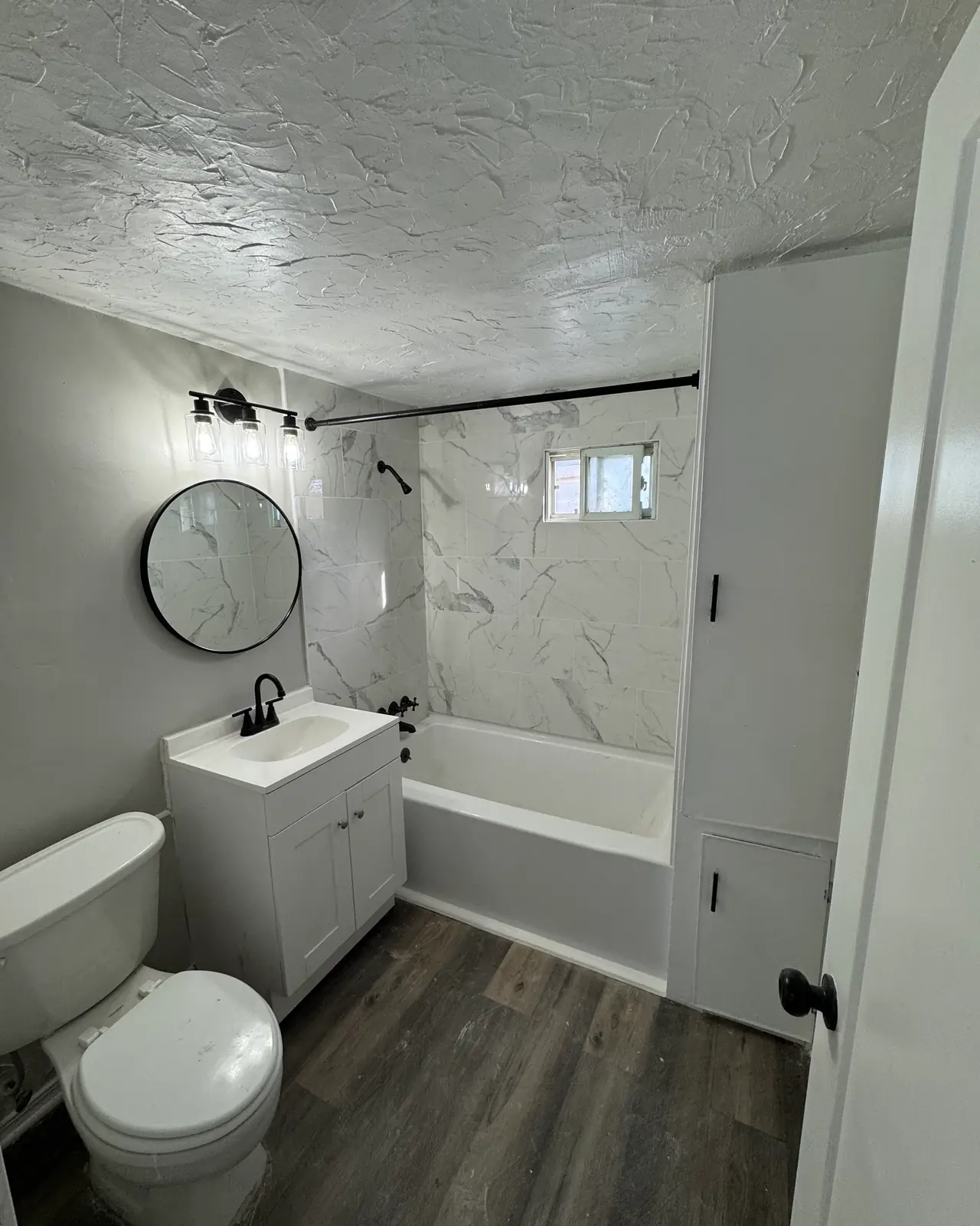 Bathroom with marble-look tub surround, white vanity, round mirror and black fixtures, after
